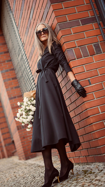Modelka w czarnej włenianej sukience zaqanna opiera się o budynek. Widać całą jej sylwetkę.